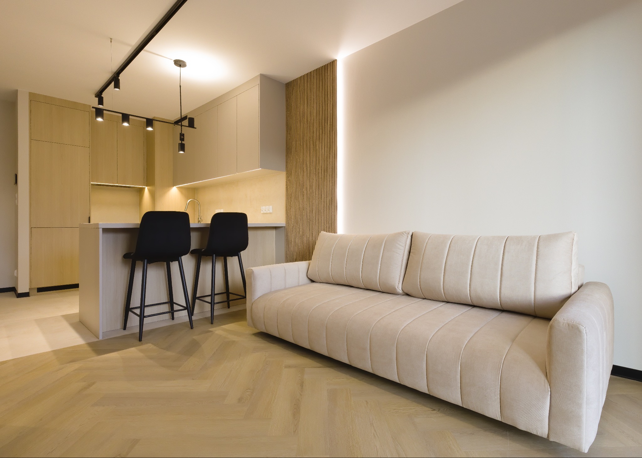 Jasne wnętrze: beżowa sofa na parkiecie w jodełkę, wyspa kuchenna z czarnymi hokerami, oświetlenie szynowe i dekoracyjna ściana z lameli. Minimalistyczny design.