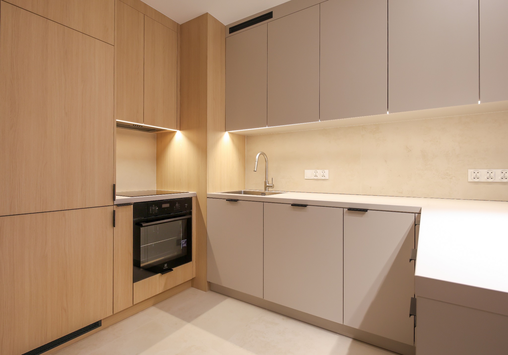 Nowoczesna kuchnia z drewnianymi i szarymi szafkami, oświetleniem LED pod szafkami wiszącymi i sprzętem AGD w zabudowie. Minimalistyczny design, jasne wnętrze.