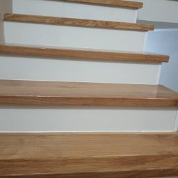 FENIKS WYKOŃCZENIA - Drewniane schody z białymi podstopnicami, widok z dołu. Polakierowane stopnie z wyraźnym usłojeniem. Górny fragment drzwi w tle.