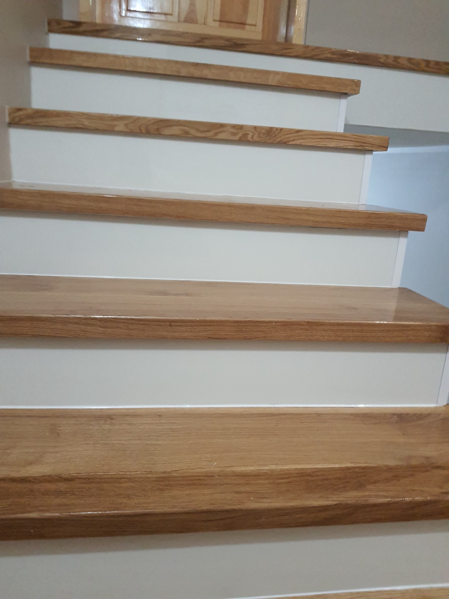 Drewniane schody z białymi podstopnicami, widok z dołu. Polakierowane stopnie z wyraźnym usłojeniem. Górny fragment drzwi w tle.