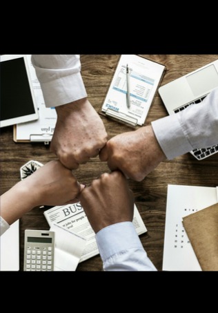 Zespół łączy siły: cztery zaciśnięte pięści na tle biurka z dokumentami, laptopem i kalkulatorem. Symbol współpracy i sukcesu w biznesie.