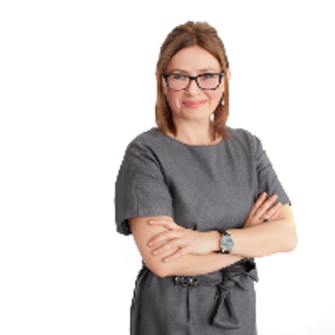 Portret kobiety w okularach, z założonymi rękami, w szarej sukience na białym tle. Profesjonalny wizerunek.