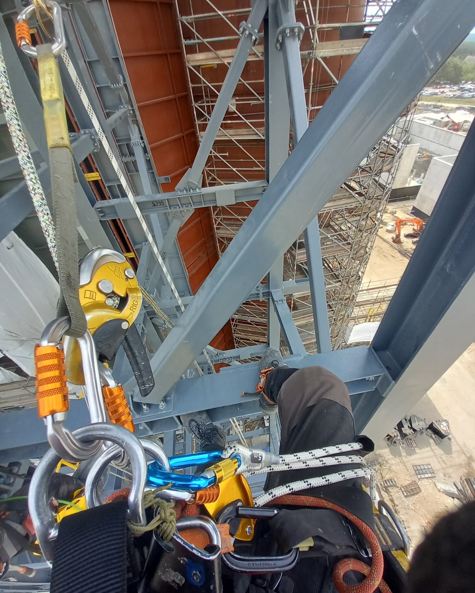 Montaż konstrukcji stalowej z perspektywy pracownika wysokościowego, widoczne liny, karabinki i elementy rusztowania w tle. Ujęcie z góry na plac budowy.