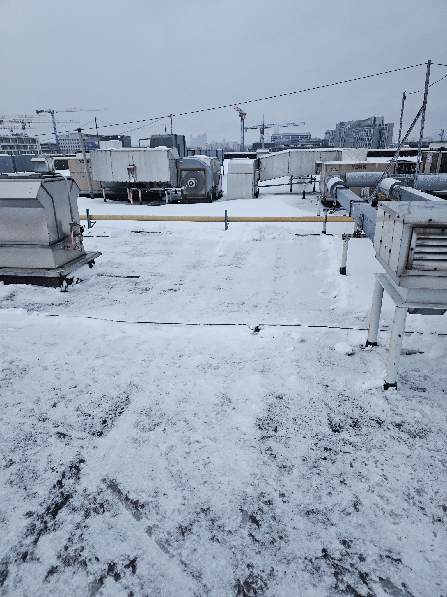 Śnieg na dachu budynku z instalacjami wentylacyjnymi i klimatyzacyjnymi. W tle widoczne budynki i żurawie budowlane w pochmurny dzień.