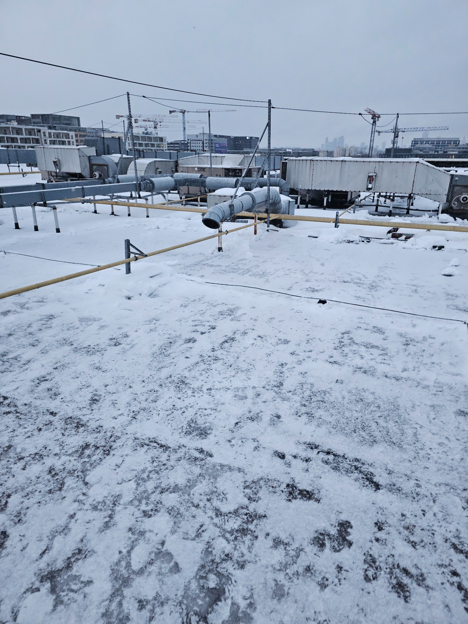 Śnieżny dach z instalacją wentylacyjną i widokiem na miasto z żurawiami w tle. Zimowy krajobraz miejski z perspektywy dachu.