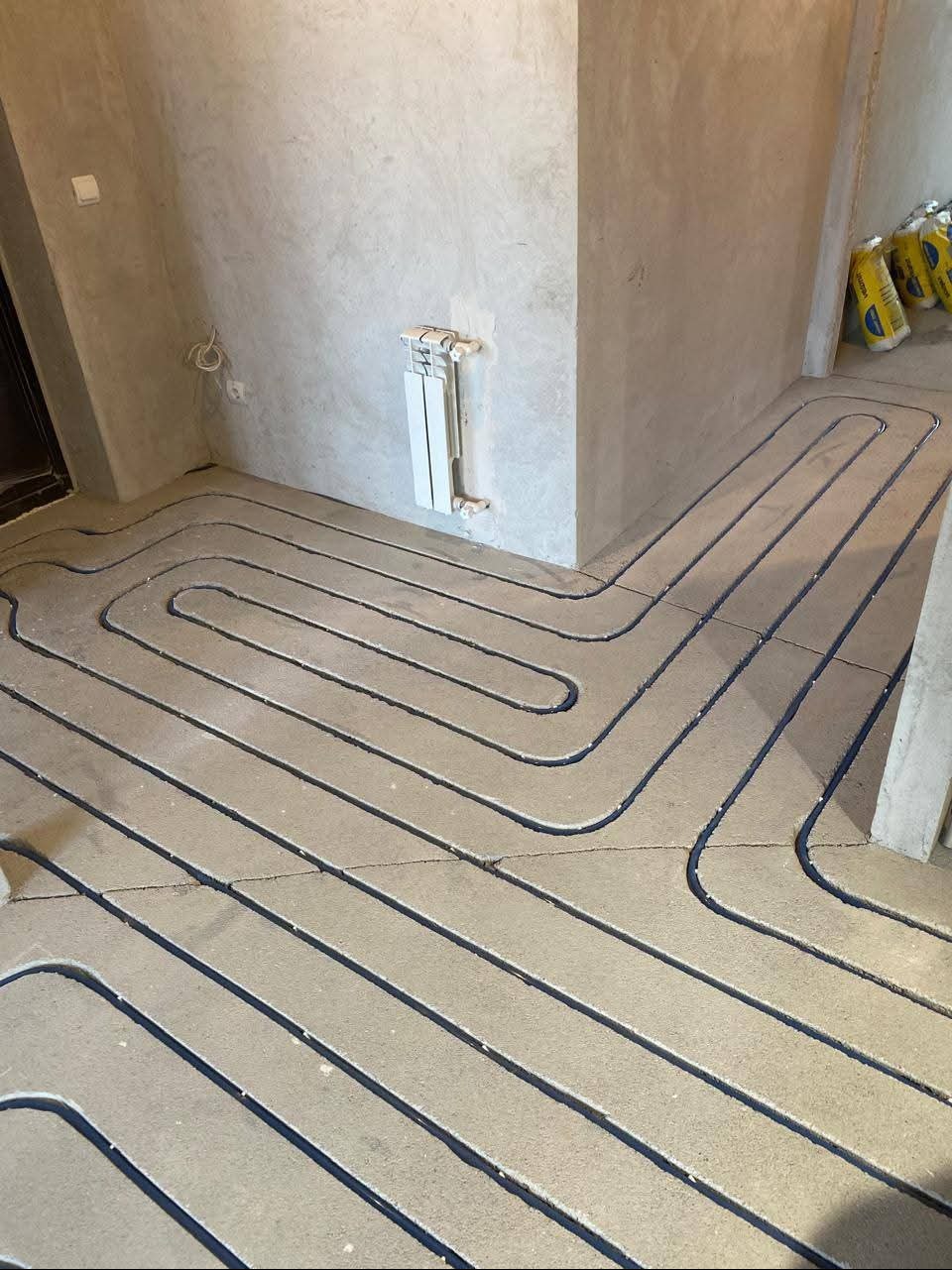 Szare rury ogrzewania podłogowego ułożone w jastrychu, widoczne przy ścianie grzejnik i worki z zaprawą. Ujęcie z góry, surowy stan wykończenia.