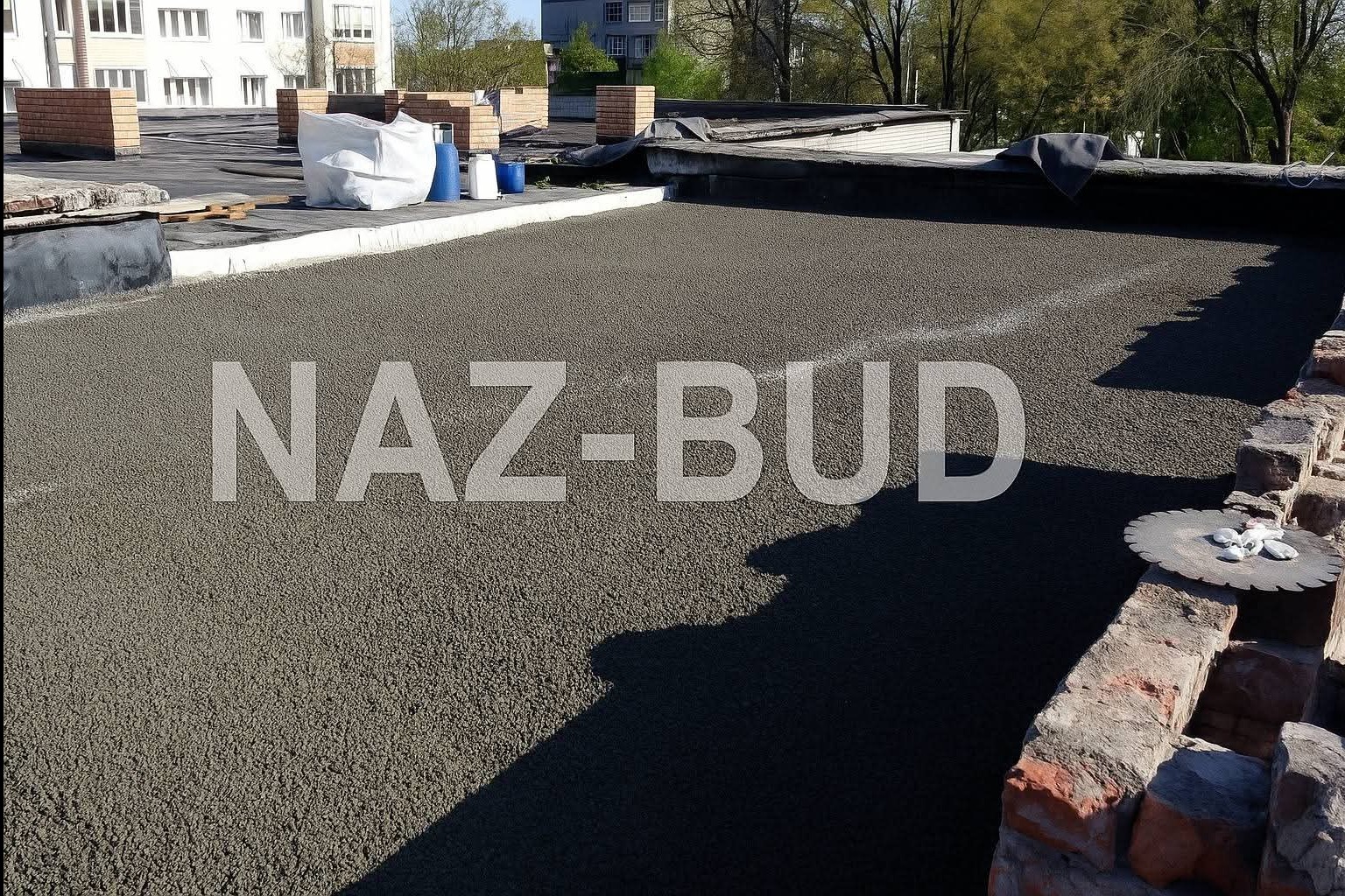 Świeżo wylana, szara posadzka na dachu budynku, z widocznym logo firmy 'NAZ-BUD' na powierzchni. W tle ceglane kominy i budynki mieszkalne.