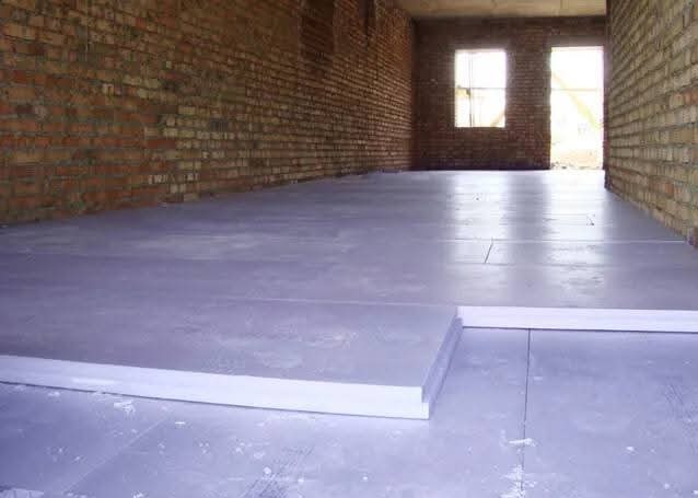 Jasnoszara, gładka powierzchnia podłogi w surowym wnętrzu z ceglanymi ścianami. Widoczne białe listwy przypodłogowe i fragmenty izolacji. Dwa okna w głębi pomieszczenia.