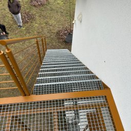 RUSLAN SOKHACH ETERNAL - Zewnętrzne schody metalowe z kratownicowymi stopniami i żółtą balustradą, prowadzące w górę wzdłuż białej ściany budynku. Widok z góry na konstrukcję i otoczenie.