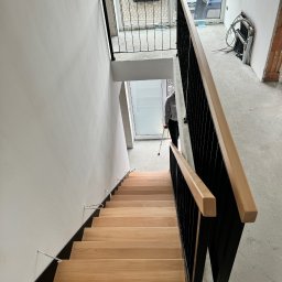 RUSLAN SOKHACH ETERNAL - Drewniane schody z czarną, metalową balustradą i jasnym drewnianym poręczą w surowym wnętrzu. Widok z góry na dół, osoba z laską w tle.