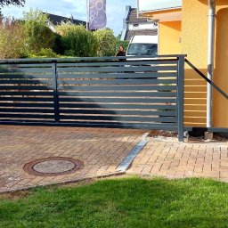 RUSLAN SOKHACH ETERNAL - Nowoczesna, przesuwna brama wjazdowa z poziomymi panelami, pomalowana na grafitowo. Brama zamontowana na posesji z kostką brukową i zielenią. W tle budynek z żółtą elewacją.