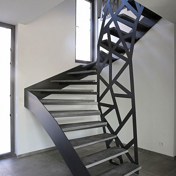 Nowoczesne schody spiralne z metalową balustradą o geometrycznym wzorze, betonowe stopnie, minimalistyczne wnętrze z oknem. Ujęcie z dołu, podkreślające design.