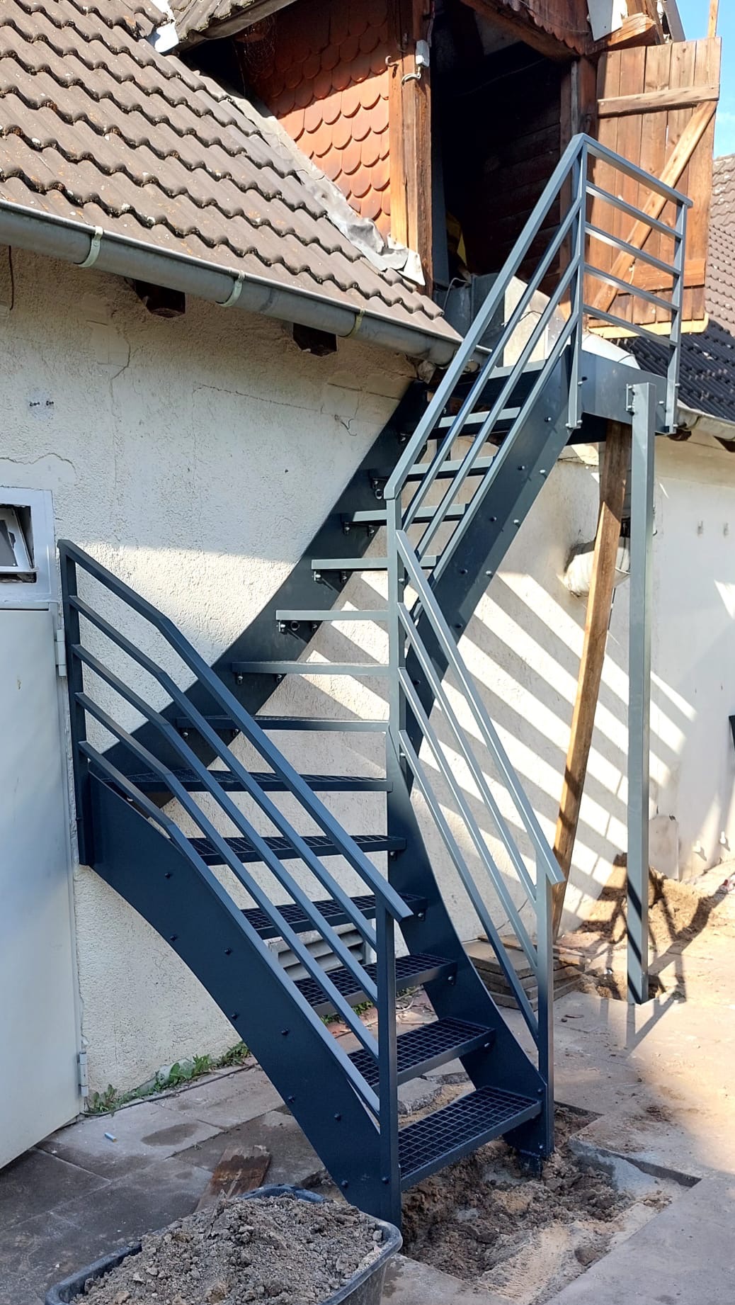 Zewnętrzne schody metalowe z balustradą, prowadzące do budynku z drewnianymi drzwiami na poddaszu. Widoczne ślady prac montażowych.