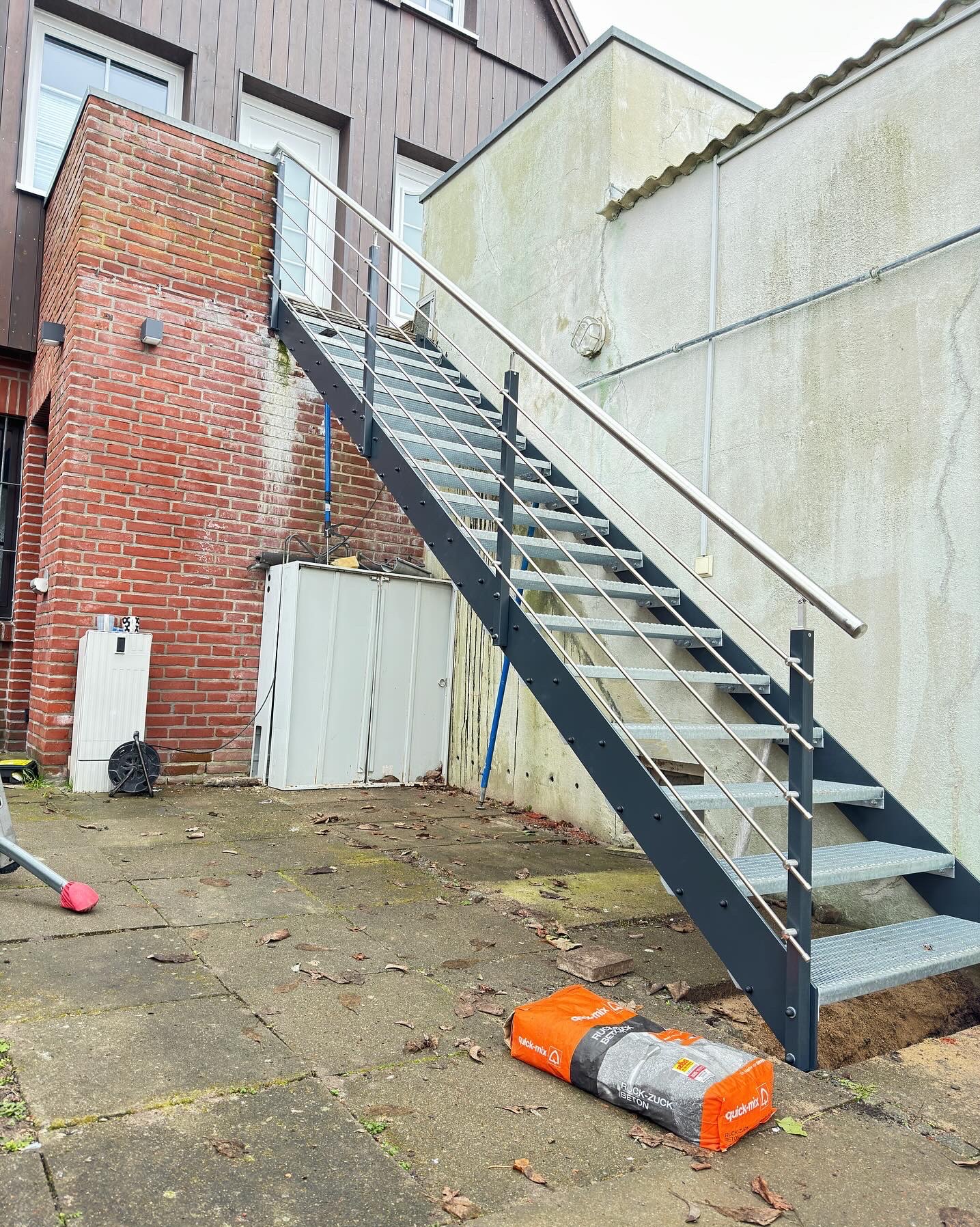 Zewnętrzne schody metalowe z poręczą ze stali nierdzewnej, prowadzące do drzwi na piętrze. Widoczny worek z betonem, sugerujący montaż. Ujęcie z dołu, podkreślające konstrukcję.