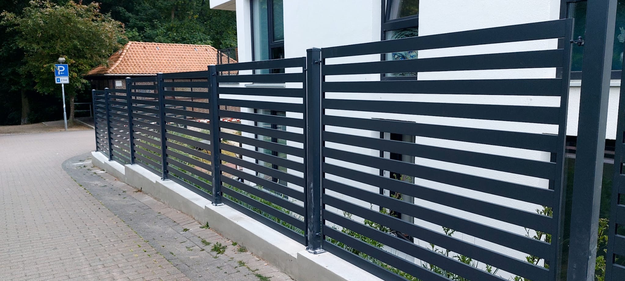 Nowoczesne ogrodzenie metalowe z poziomych, grafitowych profili, zamontowane na betonowej podmurówce, na tle białego budynku z oknami. Widok z zewnątrz posesji.