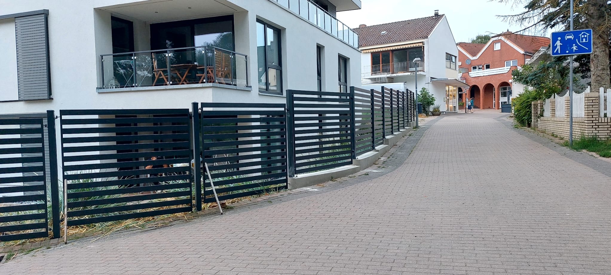 Nowoczesne, poziome ogrodzenie metalowe w kolorze antracytowym, oddzielające posesję od chodnika. W tle budynek mieszkalny z balkonem i szklaną balustradą.