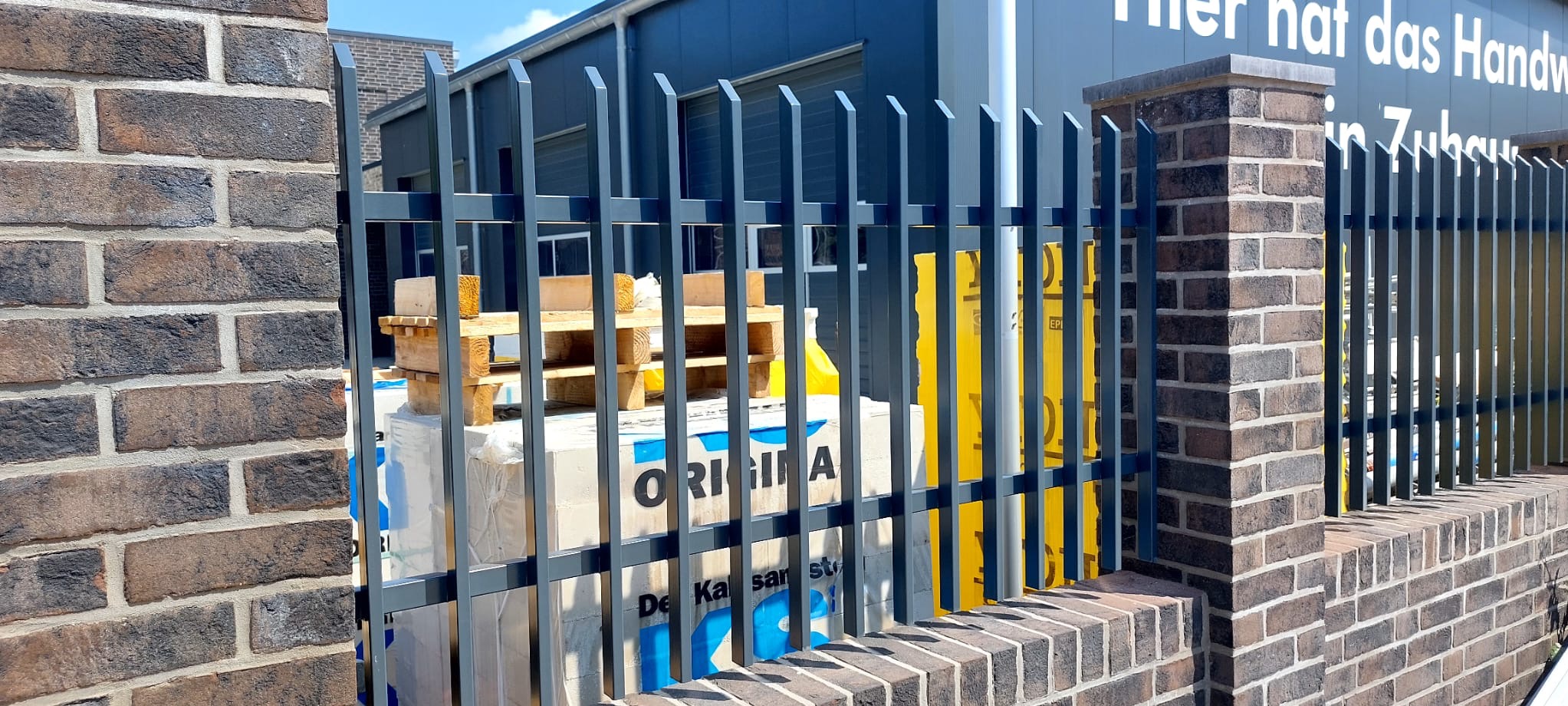 Nowoczesne ogrodzenie metalowe z pionowymi prętami, częściowo zasłaniające materiały budowlane na paletach. Ceglany murek stanowi podstawę ogrodzenia. Widok z zewnątrz posesji.