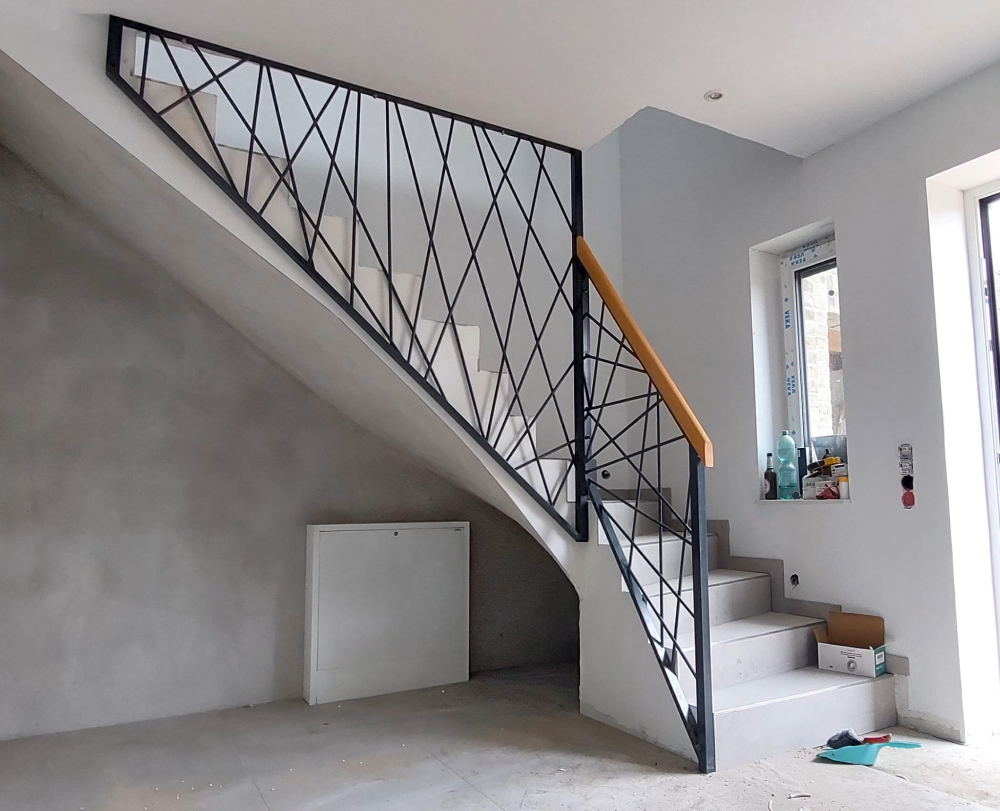 Nowoczesne schody z czarną, geometryczną balustradą i drewnianym poręczą. Widok z dołu, surowy stan wykończenia wnętrza. Minimalistyczny design.