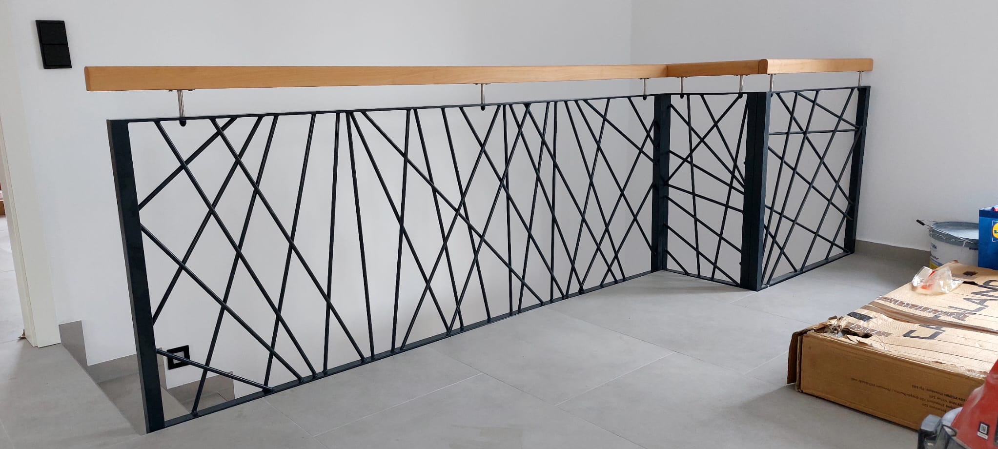 Nowoczesna balustrada wewnętrzna z czarnego metalu o geometrycznym wzorze, zwieńczona drewnianym elementem, na tle białej ściany i szarej podłogi.