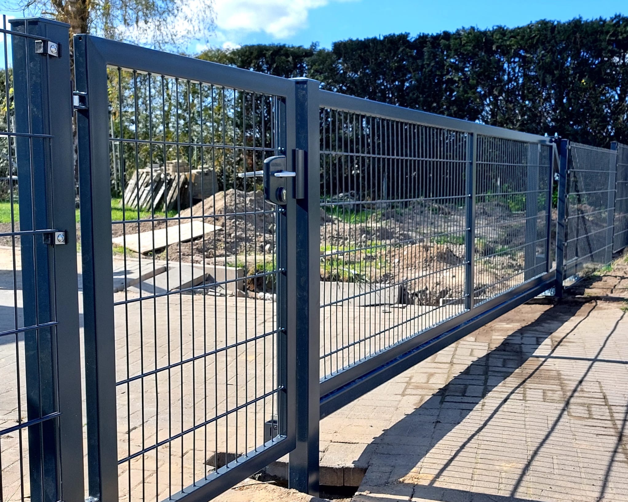 Nowoczesna brama przesuwna z metalowej siatki w Kostrzynie nad Odrą. Widoczny zamek i solidna konstrukcja. Tło: zielony żywopłot i przygotowany teren pod dalsze prace.