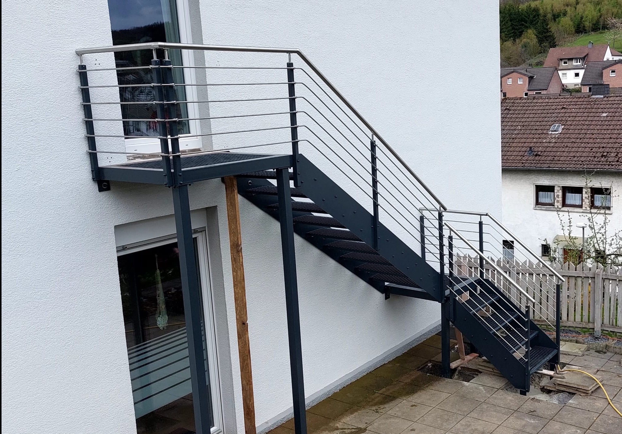 Zewnętrzne schody metalowe z balustradą ze stali nierdzewnej, prowadzące na balkon. Konstrukcja przymocowana do białej ściany budynku. Widok z dołu.