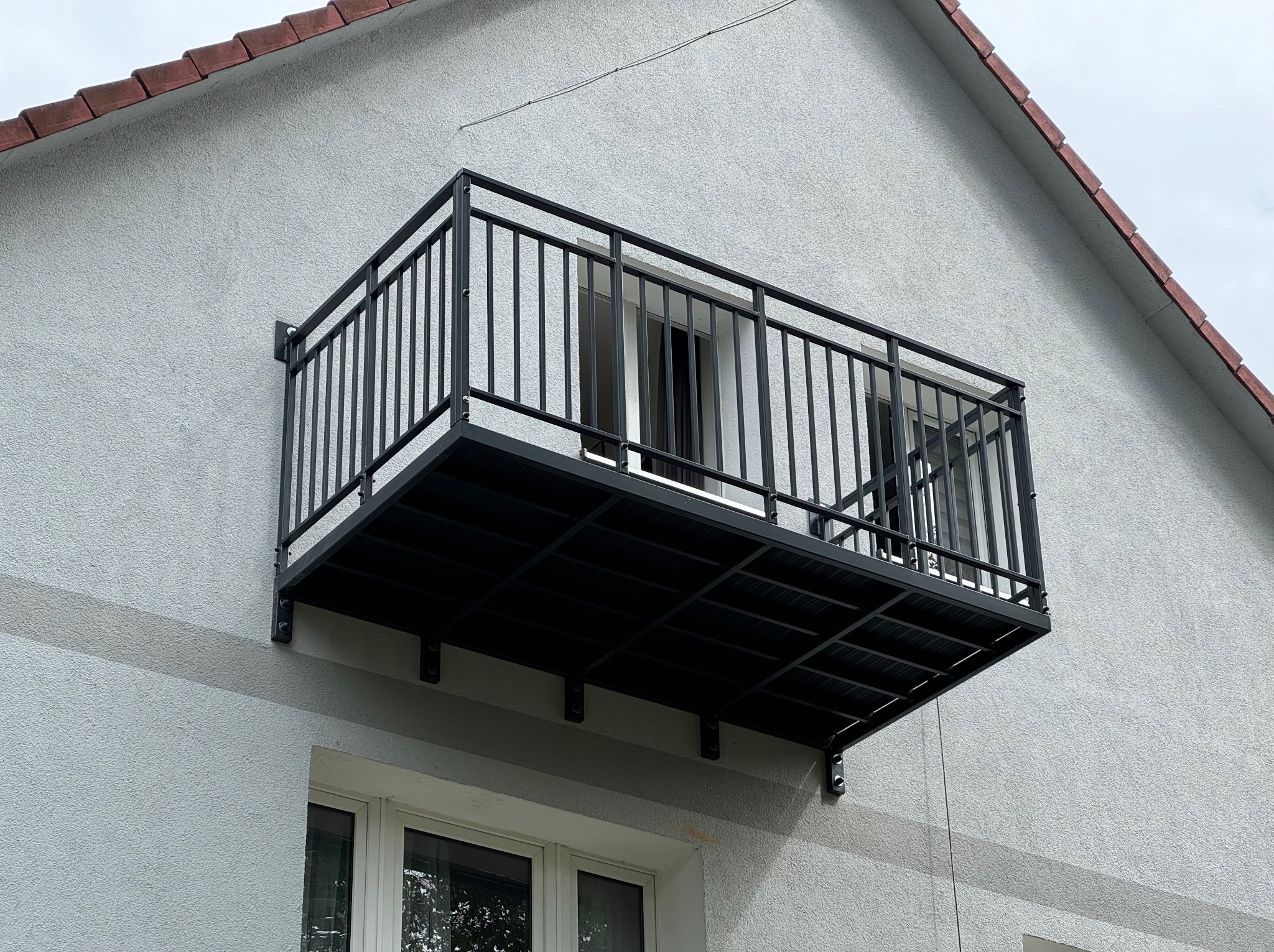 Czarny, metalowy balkon z barierką, zamontowany na elewacji budynku. Widoczne okna i fragment dachu. Minimalistyczny design, proste linie, szara elewacja.