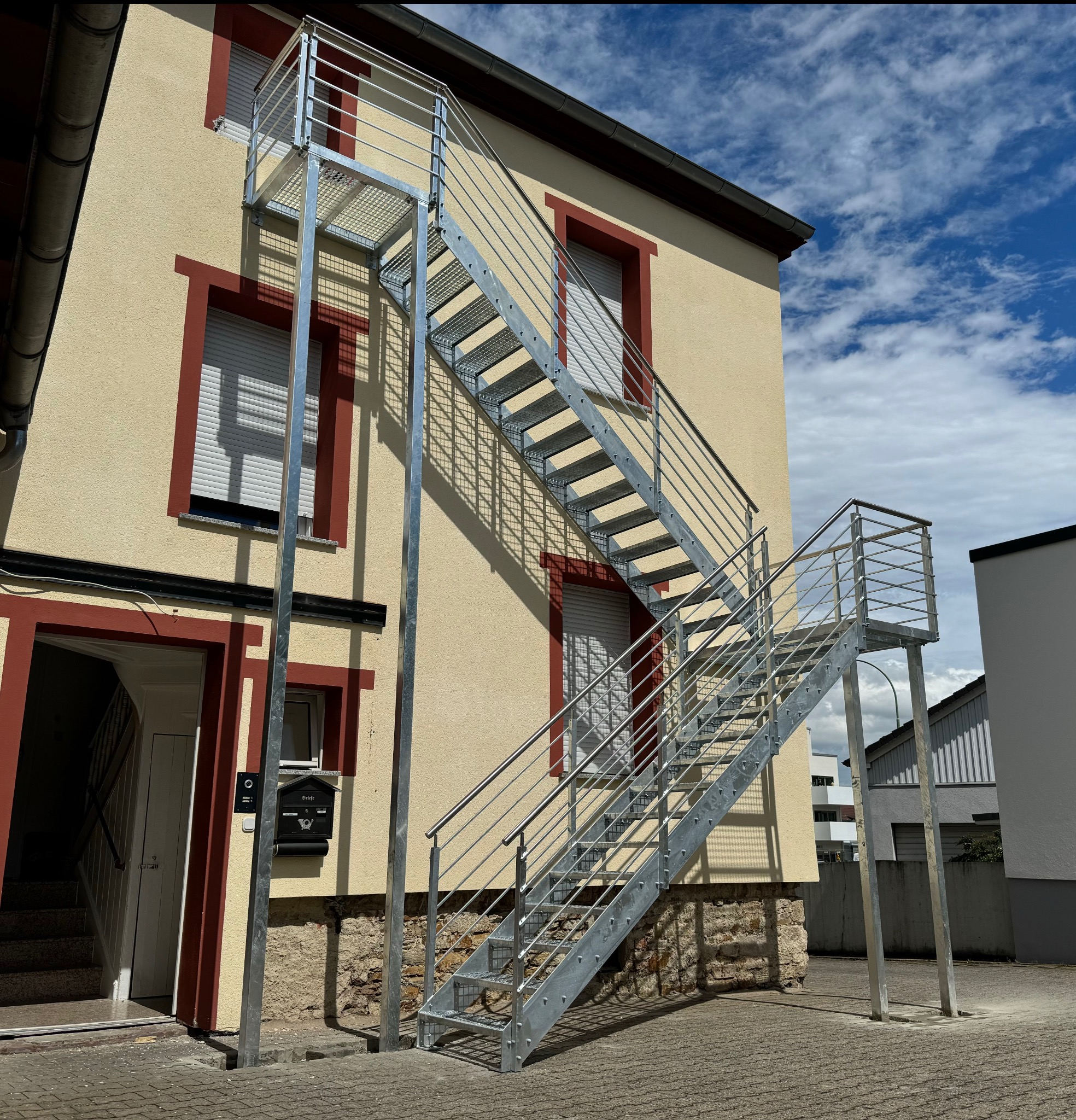 Zewnętrzne, metalowe schody z balustradą, prowadzące na wyższą kondygnację budynku o beżowej elewacji z czerwonymi obramowaniami okien. Widok z dołu, błękitne niebo w tle.