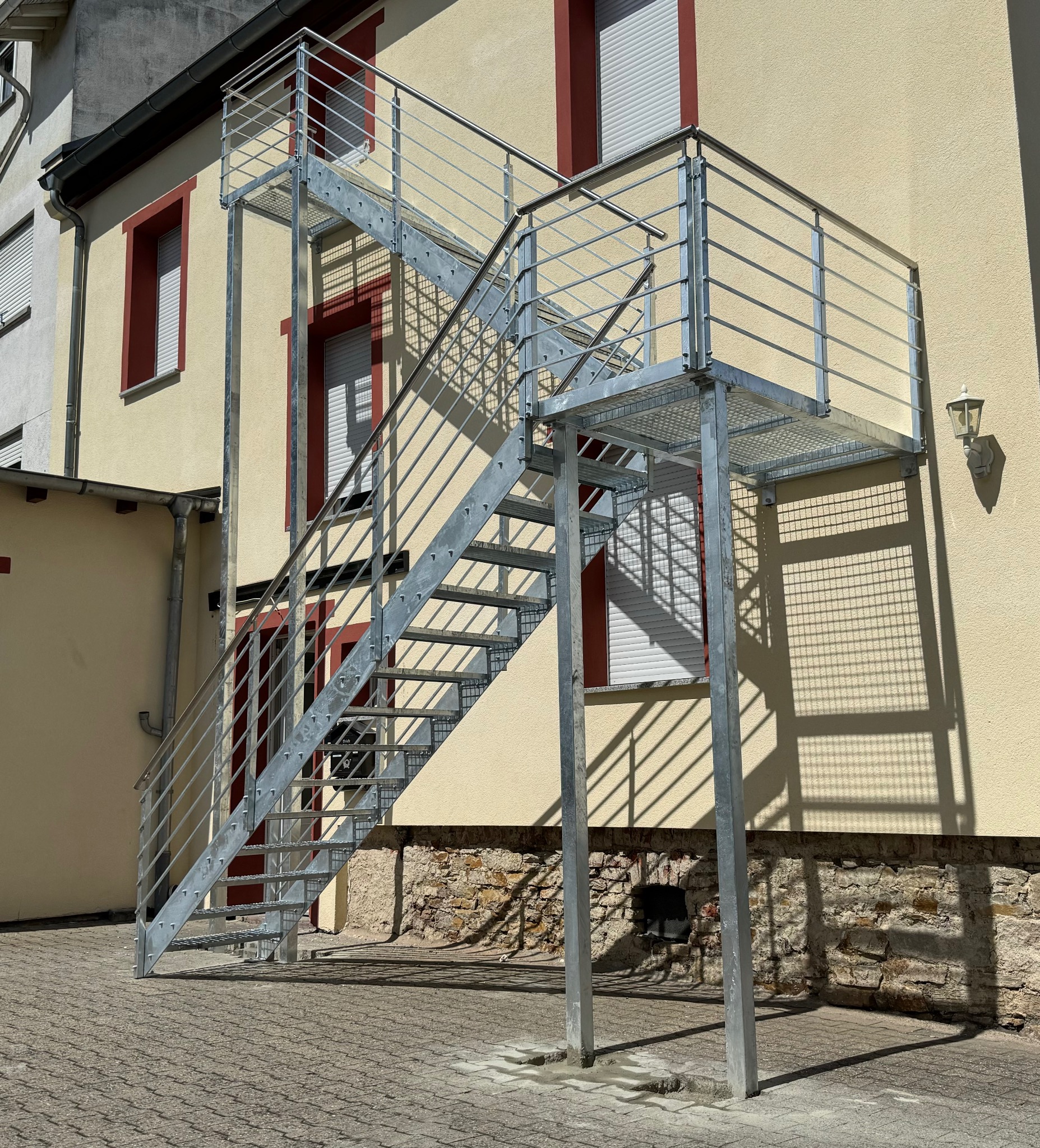 Zewnętrzne, metalowe schody z platformą, prowadzące do budynku o żółtej elewacji. Konstrukcja ze srebrnych rurek i kratownicy, rzucająca wyraźny cień na ścianę.