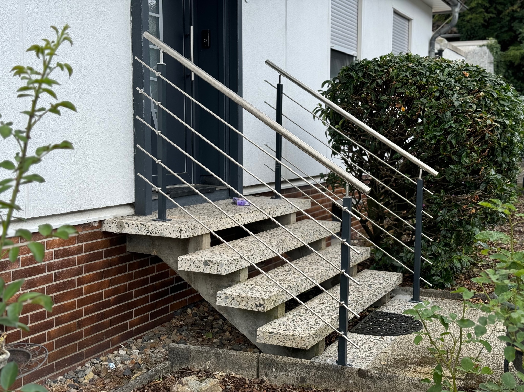 Zewnętrzne schody z lastryko z nowoczesną balustradą ze stali nierdzewnej, prowadzące do ciemnych drzwi wejściowych. Widoczne elementy architektury budynku i roślinność.