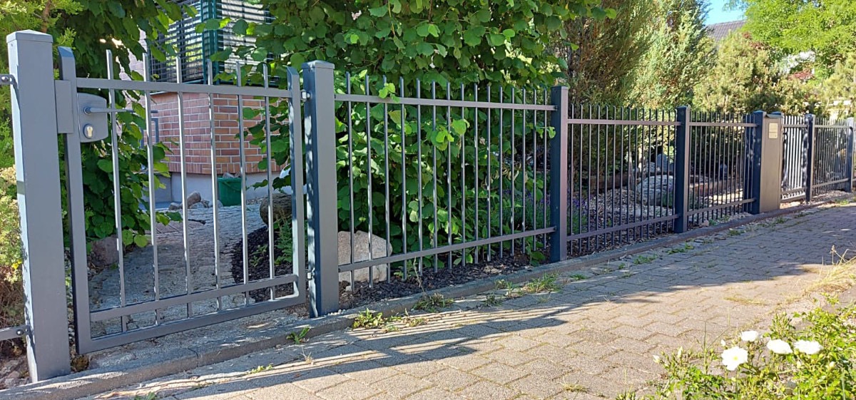 Nowoczesne, szare ogrodzenie metalowe z pionowymi prętami, zamykane furtką z klamką, otoczone zielenią i brukowaną ścieżką. Widok z zewnątrz posesji.