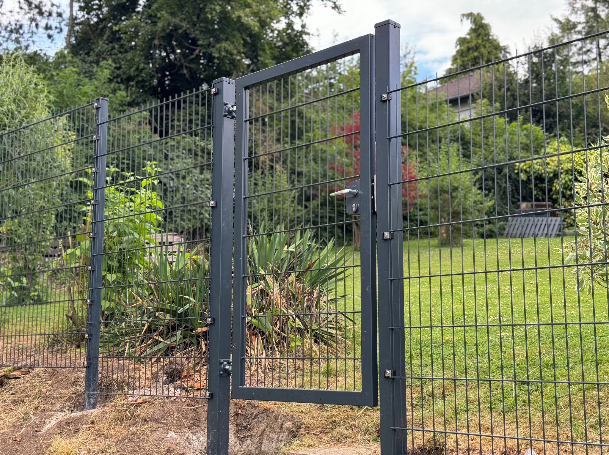 Otwarta, nowoczesna furtka z metalowej siatki na tle zieleni ogrodu. Widoczne mocowania i klamka. Minimalistyczny design, szara kolorystyka.