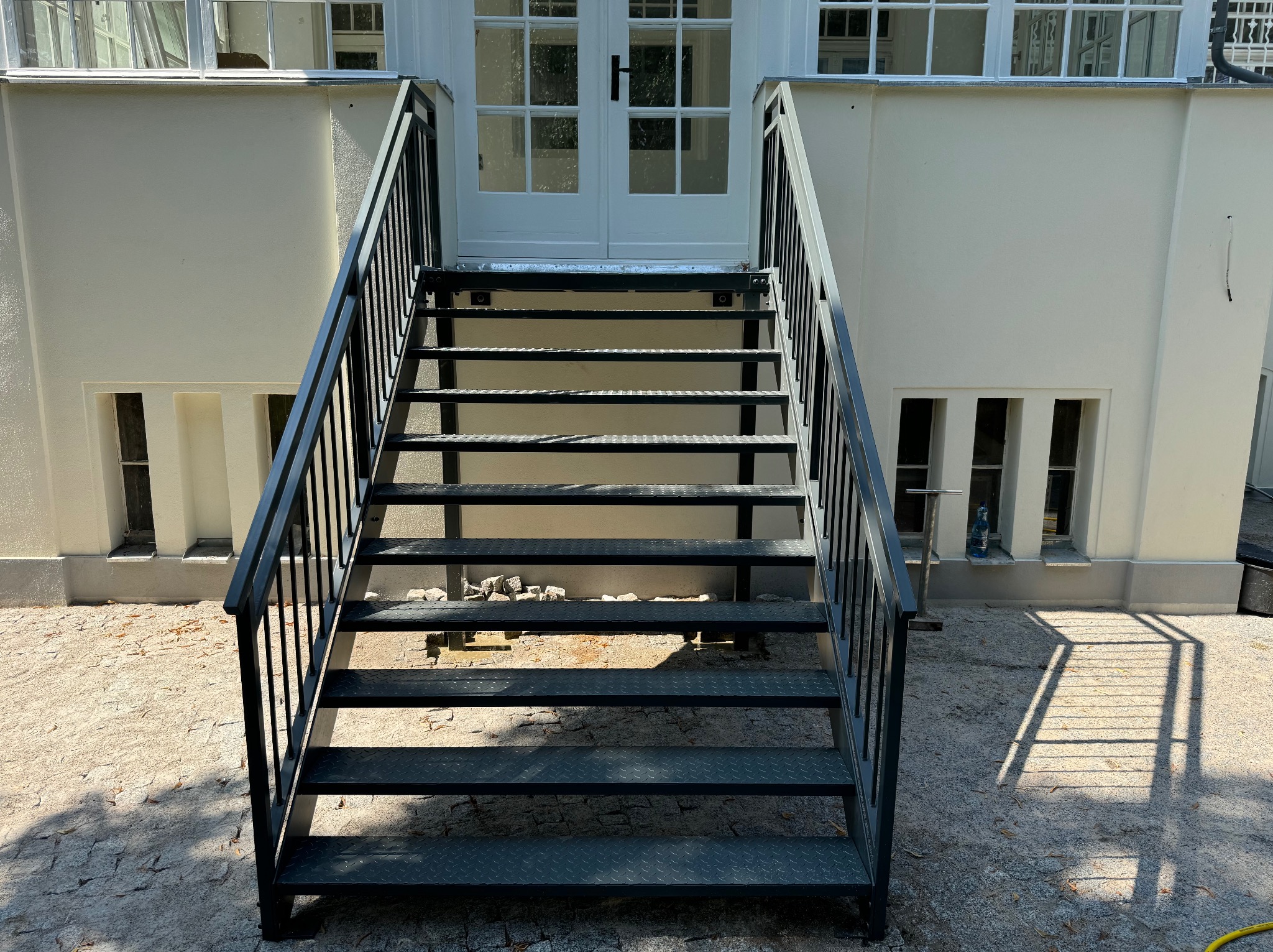 Zewnętrzne schody metalowe z czarną balustradą prowadzące do białych drzwi wejściowych. Widoczne detale konstrukcyjne i cień rzucany przez schody na podłoże.