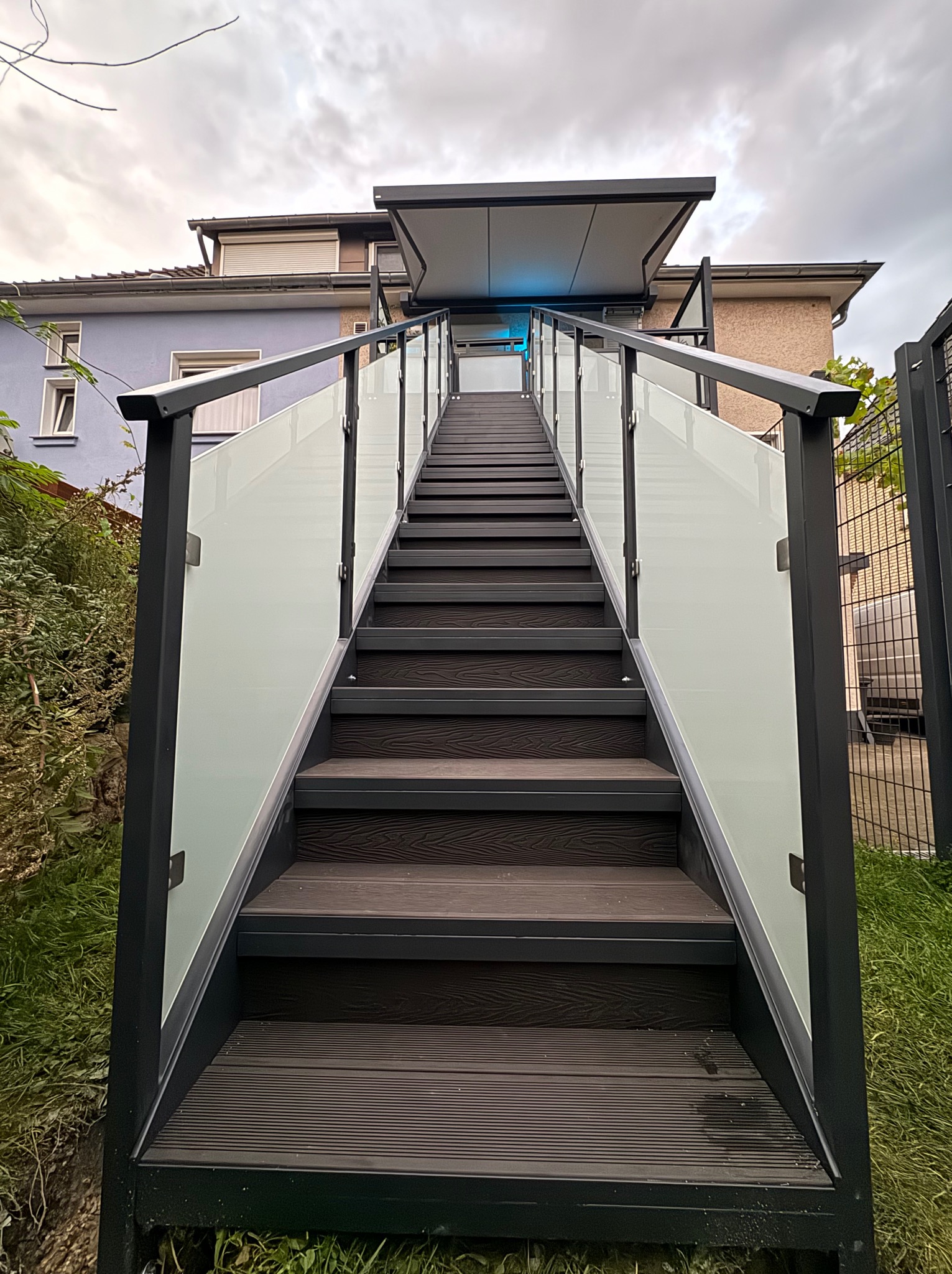 Nowoczesne schody zewnętrzne z balustradą ze szkła i metalu, prowadzące do zadaszonego wejścia. Widok z dołu, podkreślający perspektywę i solidność konstrukcji.