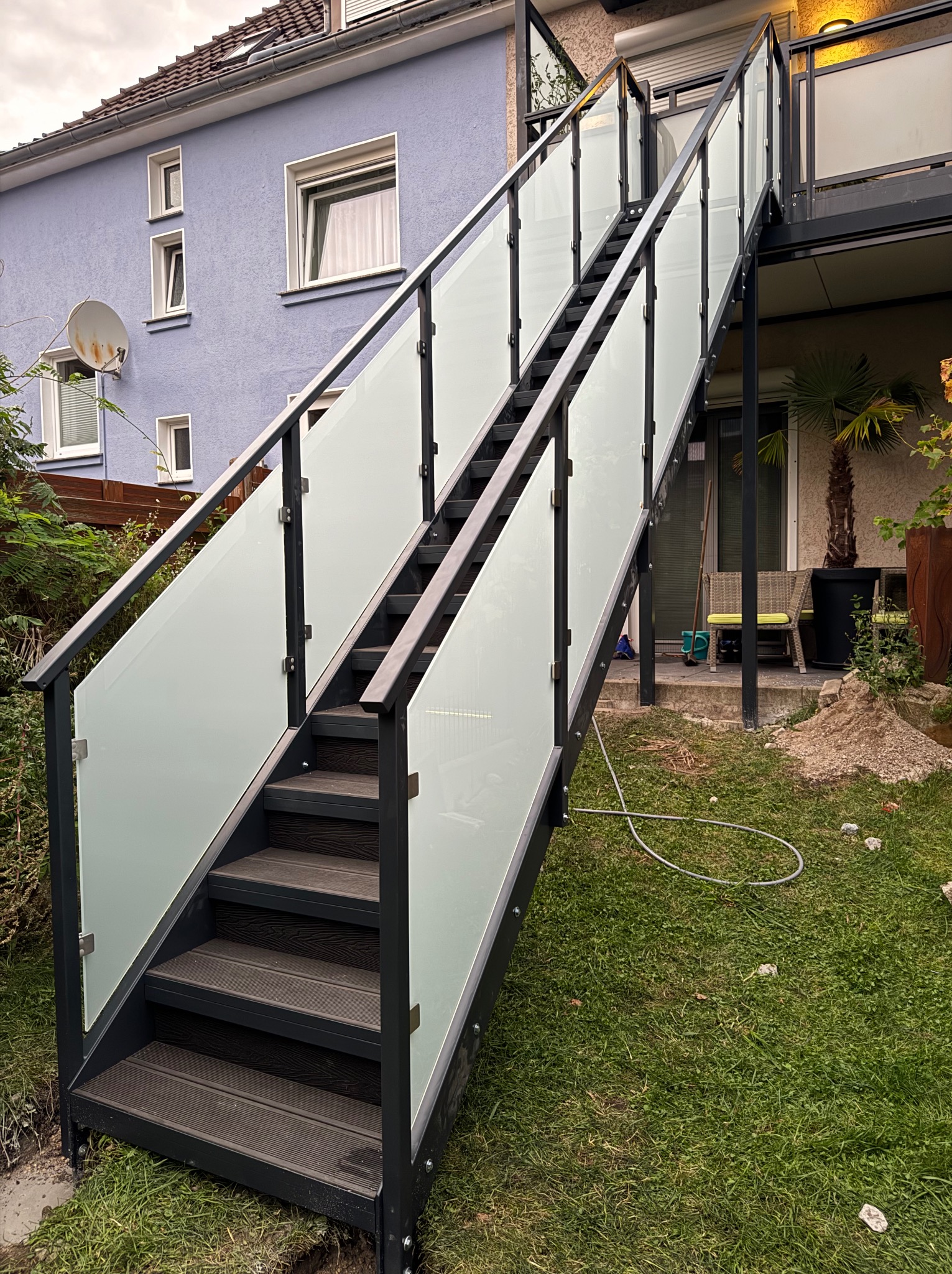 Zewnętrzne schody z balustradą ze szkła i metalu prowadzące na balkon. Widok z dołu, na tle domu i zieleni.