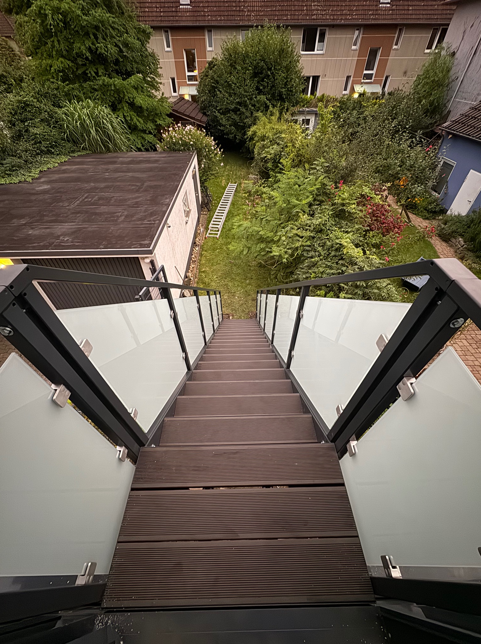 Nowoczesne schody zewnętrzne z drewnopodobnych desek z balustradą ze szkła i czarnego metalu, widok z góry na ogród i budynki w tle.