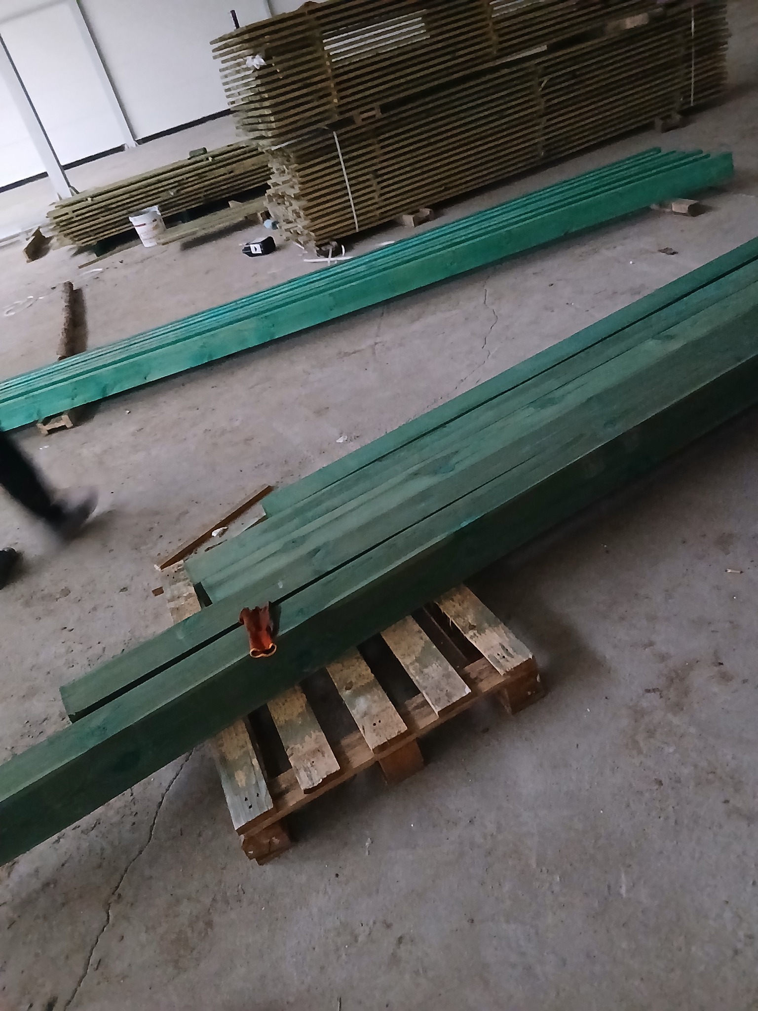 Impregnowane, zielone belki drewniane ułożone na palecie w magazynie. W tle stosy surowego drewna. Widoczny fragment nogi osoby wchodzącej w kadr.