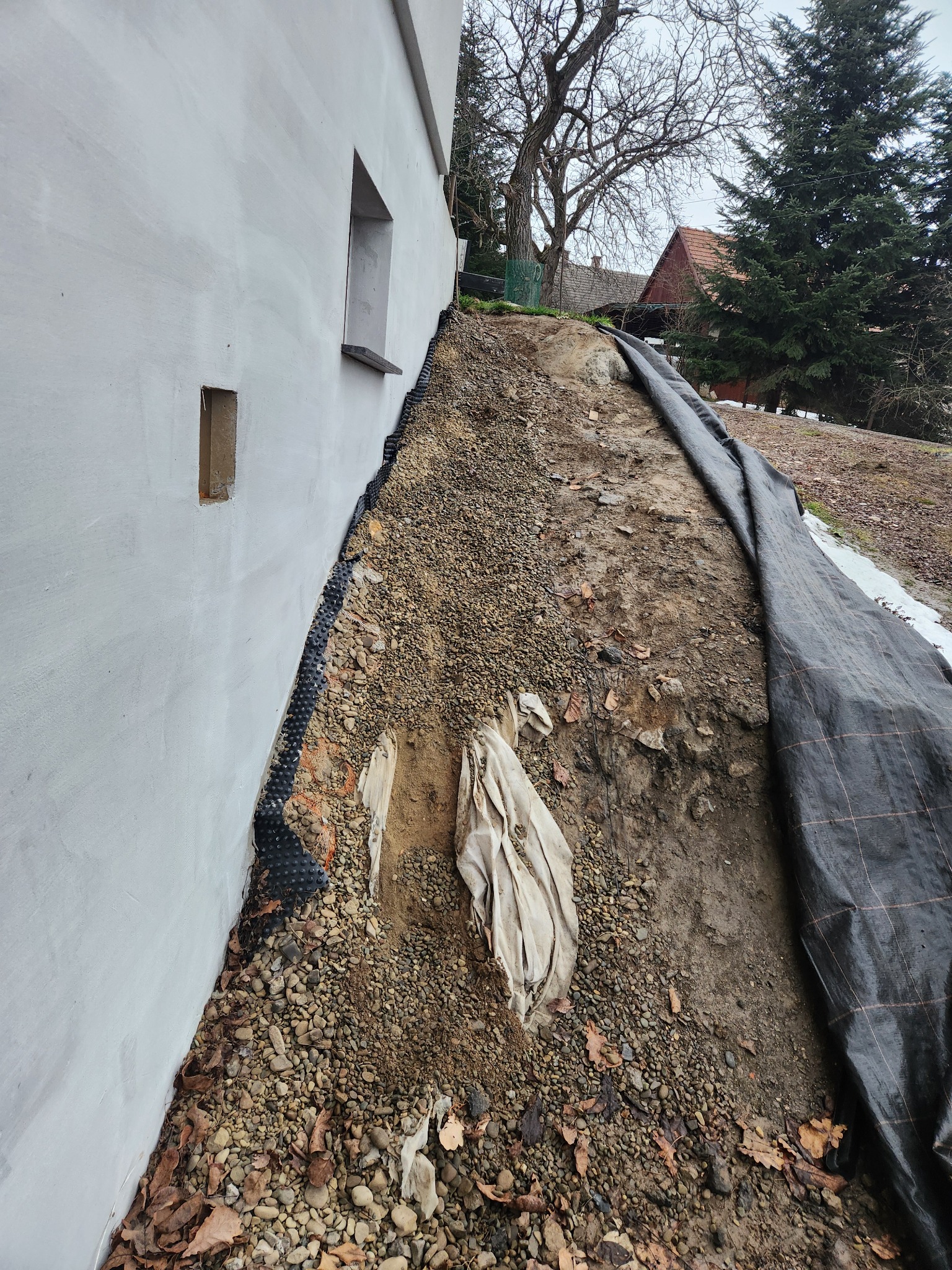 Przygotowanie terenu pod kostkę w Wieliczce: podsypka z kamienia, geowłóknina przy ścianie budynku. Widoczne narzędzia i materiały budowlane.