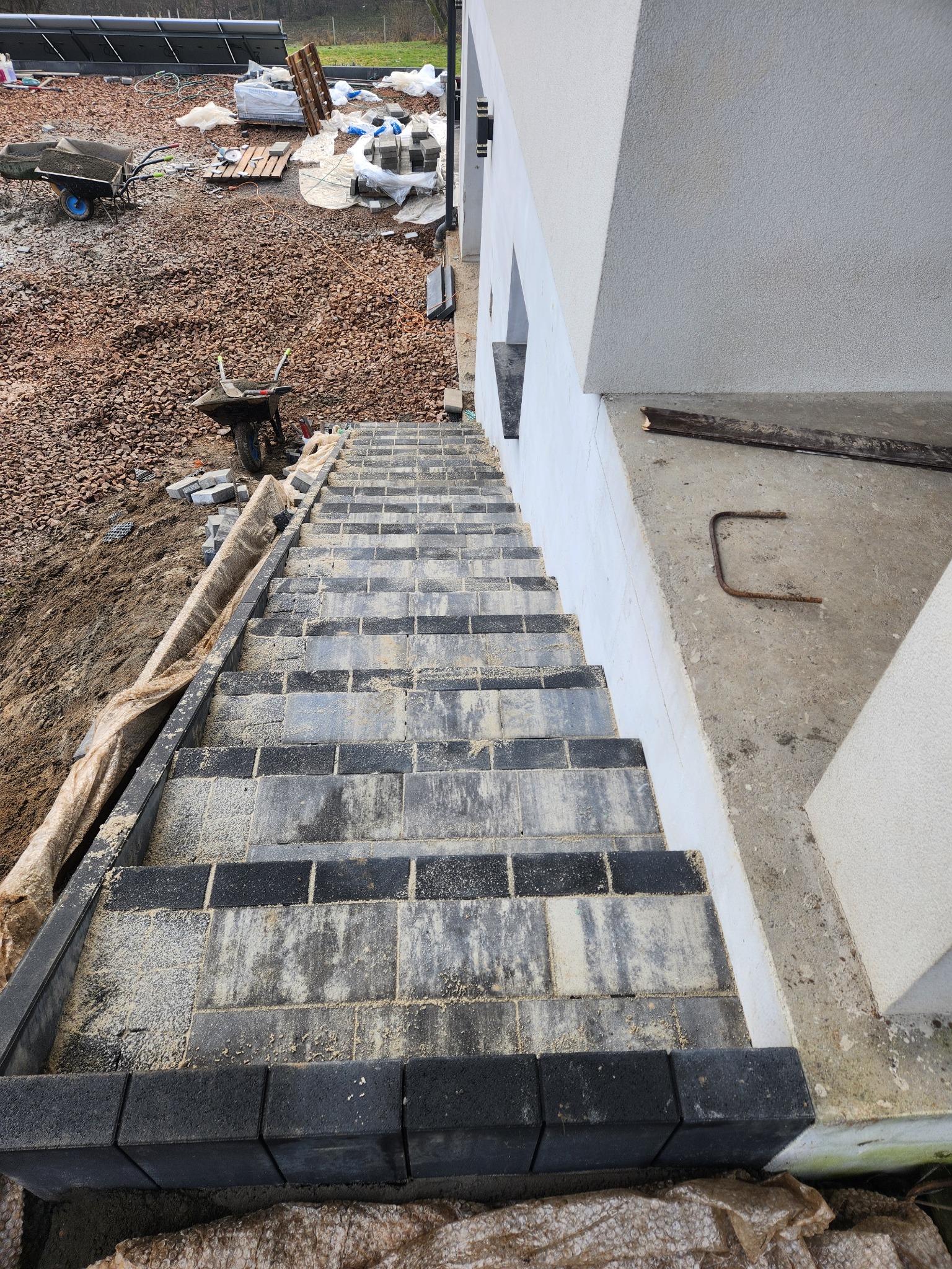 Schody z kostki brukowej w odcieniach szarości, prowadzące w dół. Widoczne narzędzia i materiały budowlane w tle. Ujęcie z góry, podkreślające geometrię i fakturę kostki.