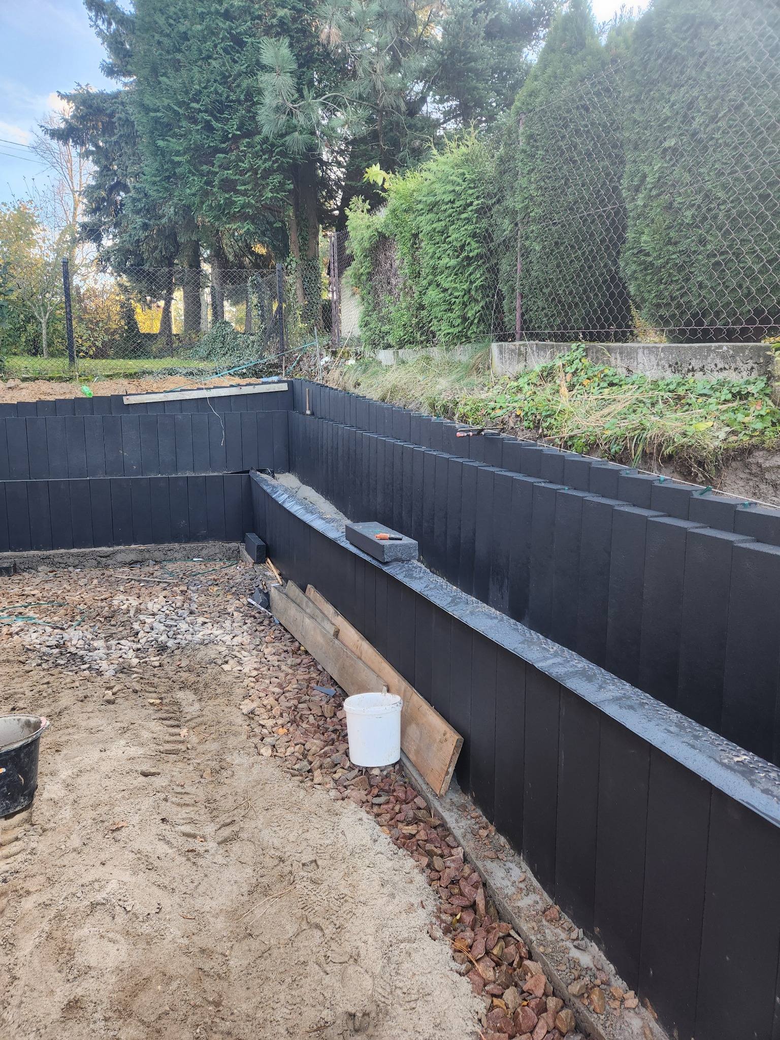 Budowa muru oporowego z czarnych bloczków w Wieliczce. Widoczne przygotowane podłoże i fragment ogrodu. Prace ziemne i konstrukcyjne w toku.