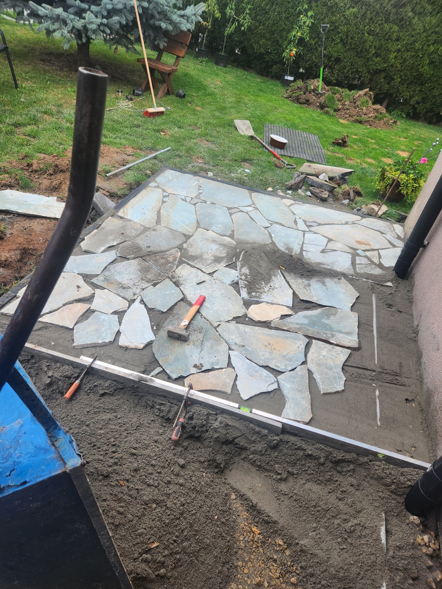 Fragment układanej ścieżki z nieregularnych kamieni na podsypce piaskowej, widoczne narzędzia i elementy otoczenia, w tle trawnik i roślinność.