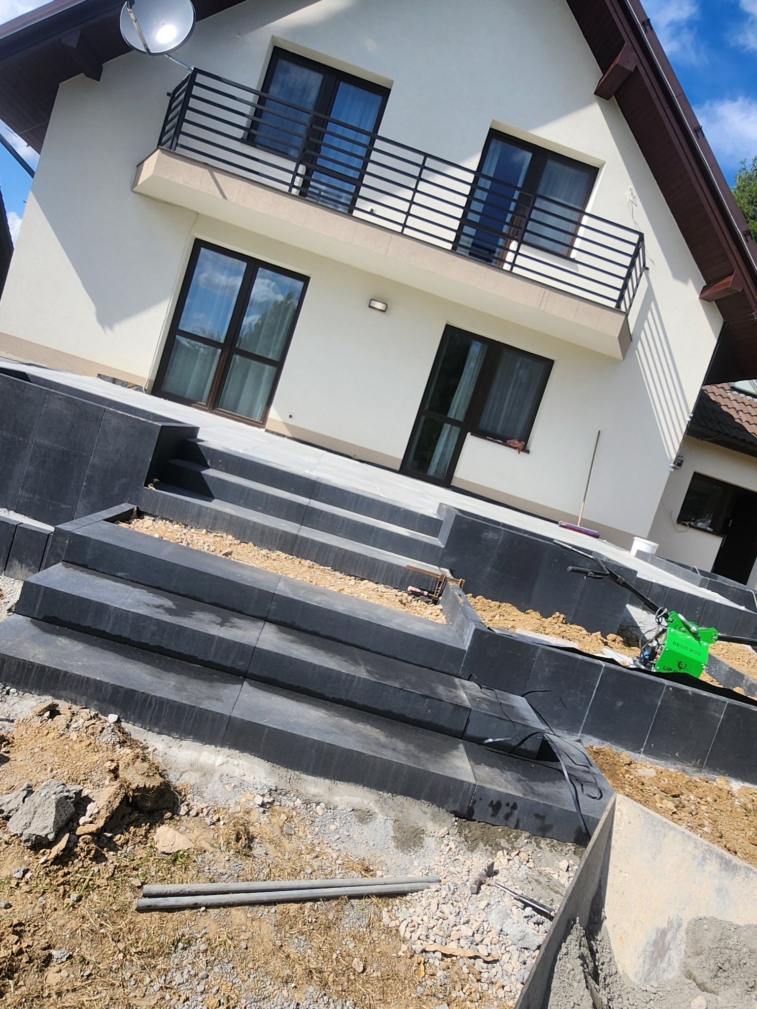 Nowoczesne schody z ciemnej kostki prowadzące do domu z balkonem. Widoczne narzędzia i materiały budowlane, sugerujące etap prac wykończeniowych.