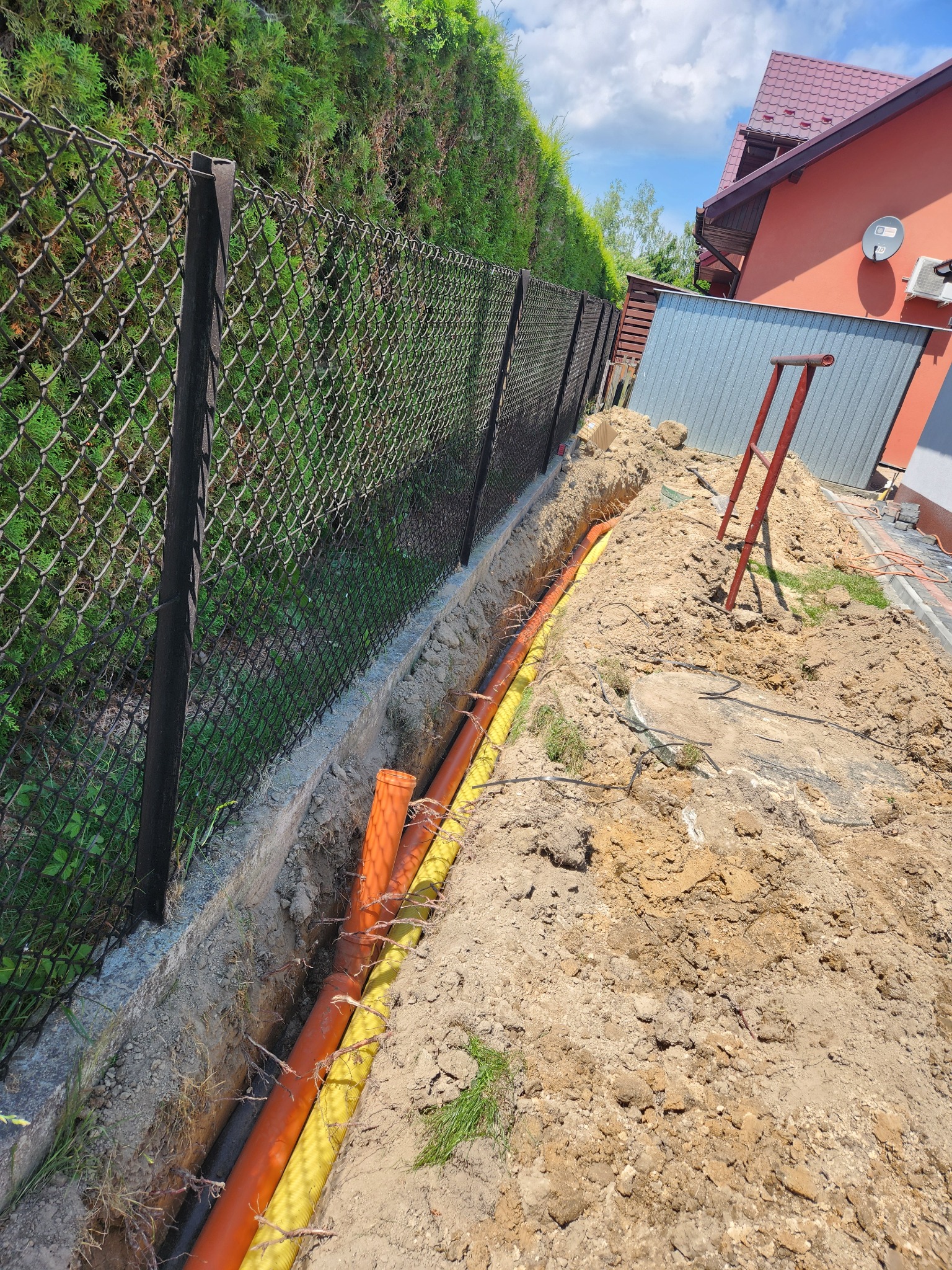 Wykop z ułożonymi rurami (żółta, pomarańczowa, czarna) wzdłuż betonowego fundamentu ogrodzenia z siatki. W tle dom z czerwoną dachówką i drabina.