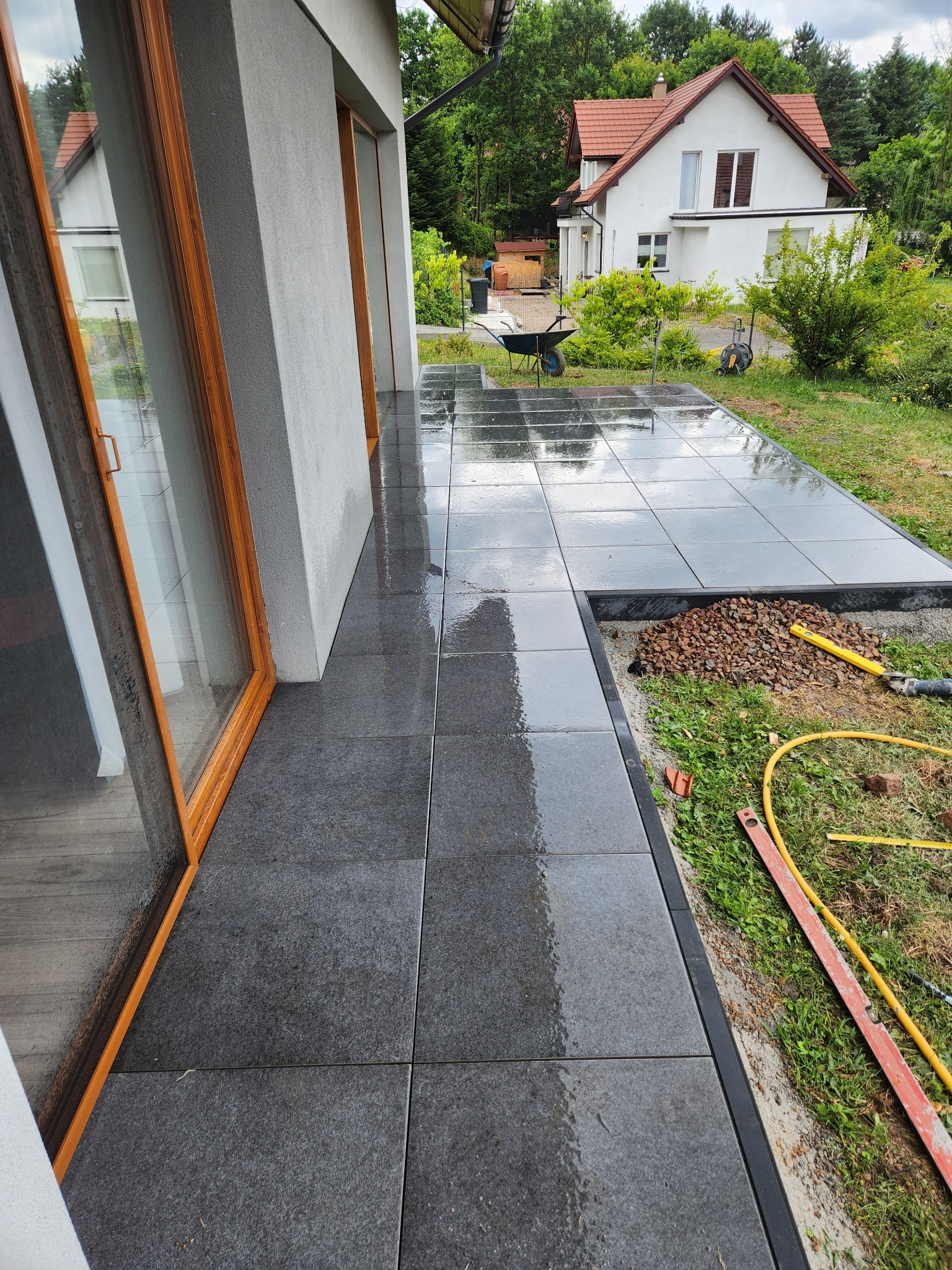 Nowoczesny taras z ciemnej kostki brukowej po deszczu, przylegający do budynku z dużym oknem. Widoczne narzędzia i materiały przygotowane do dalszych prac.