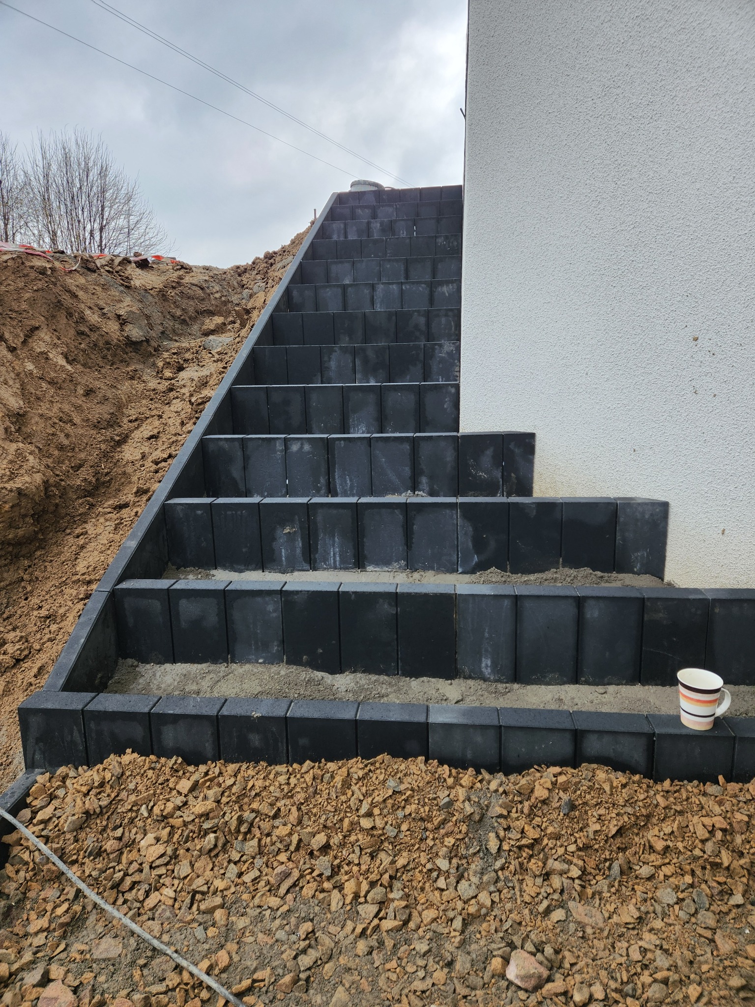 Schody z ciemnej kostki brukowej prowadzące do budynku z jasną elewacją, widoczne prace ziemne i kubek. Ujęcie z dołu, podkreślające solidność wykonania.