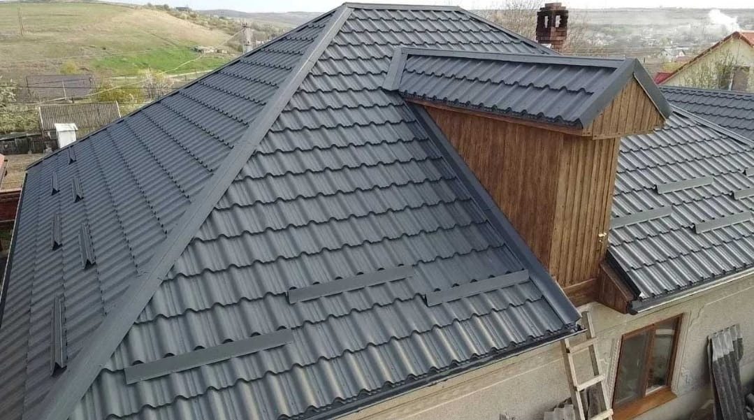 Szary dach z blachy falistej na domu jednorodzinnym, widok z góry, z kominem obłożonym drewnem. W tle pagórkowaty krajobraz.