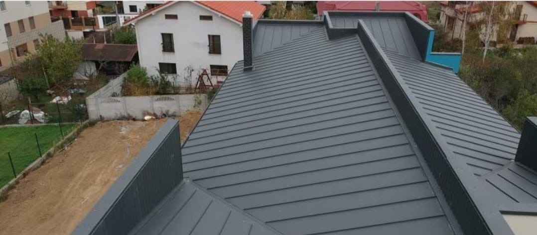 Szary, blaszany dach z kominem na tle zabudowy jednorodzinnej i zieleni. Widok z góry, prezentujący regularny układ blachy i detale wykończenia.