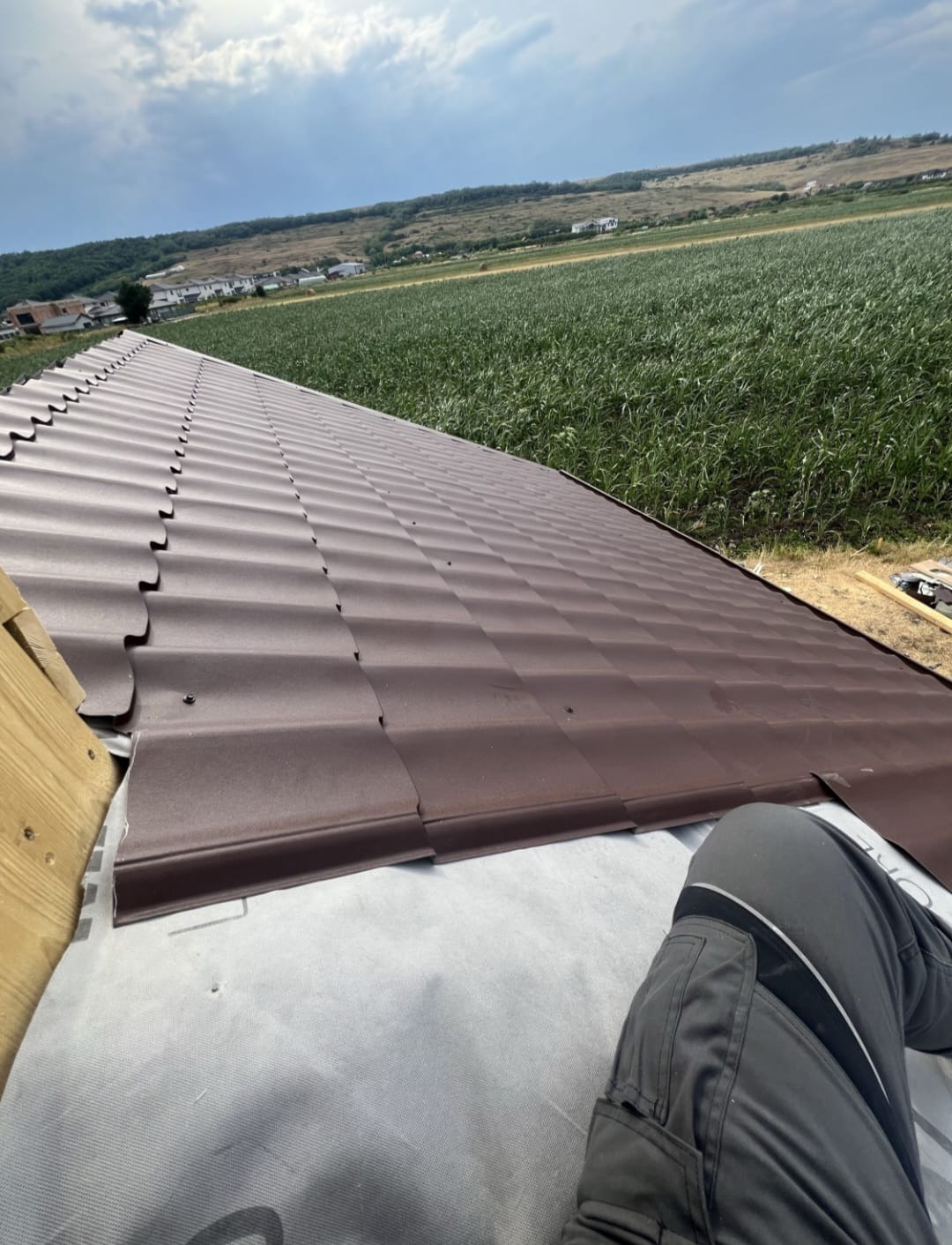 Brązowe panele dachowe ułożone na membranie, widok z góry na tle zielonych pól i zabudowań. Fragment nogi w szarych spodniach roboczych w prawym dolnym rogu.