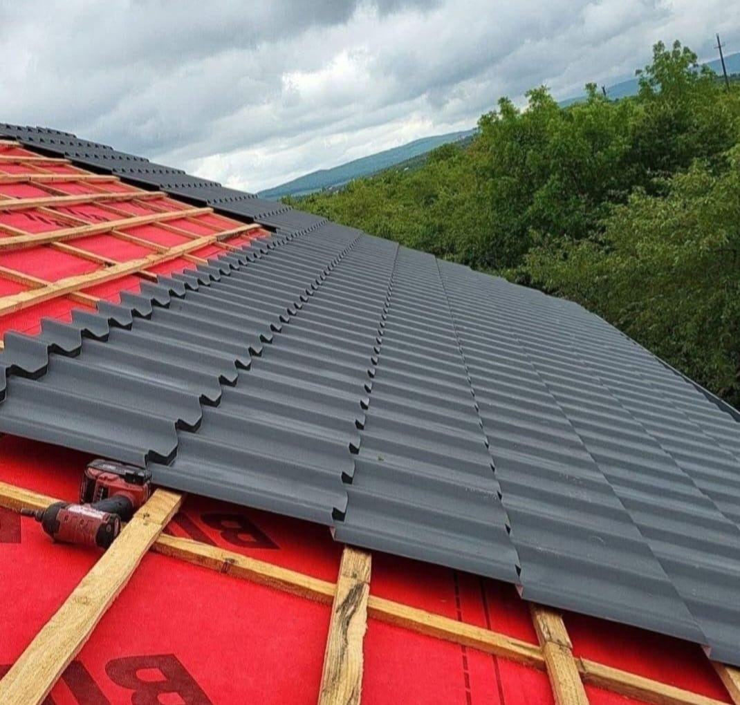 Częściowo pokryty dach blachodachówką w kolorze grafitowym, widoczna więźba i narzędzia. W tle zieleń drzew i pochmurne niebo. Etap krycia dachu.