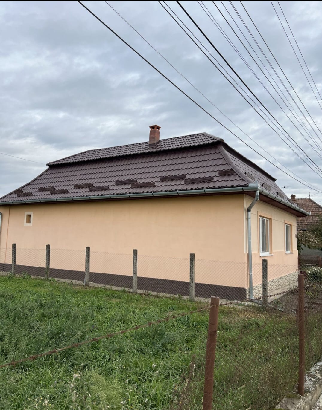 Dom jednorodzinny z brązowym dachem z blachodachówki, widoczny z boku, z rynną i kominem. Ogrodzenie z siatki i trawa na pierwszym planie. Pochmurne niebo w tle.