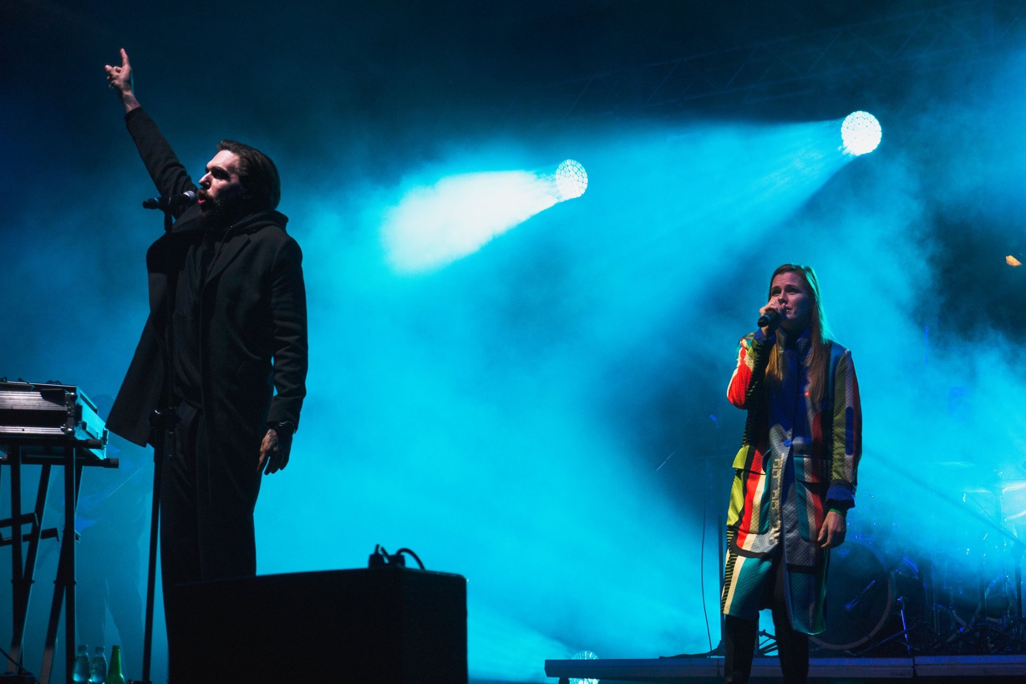 Duet muzyczny na scenie w niebieskim oświetleniu. Mężczyzna w czarnym płaszczu śpiewa do mikrofonu, obok kobieta w kolorowym płaszczu również śpiewa. Widoczne instrumenty i sprzęt nagłośnieniowy.